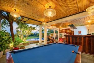 Uma mesa de bilhar com teto de madeira e luminárias pendentes, cercada por plantas em vasos e vista para uma piscina.