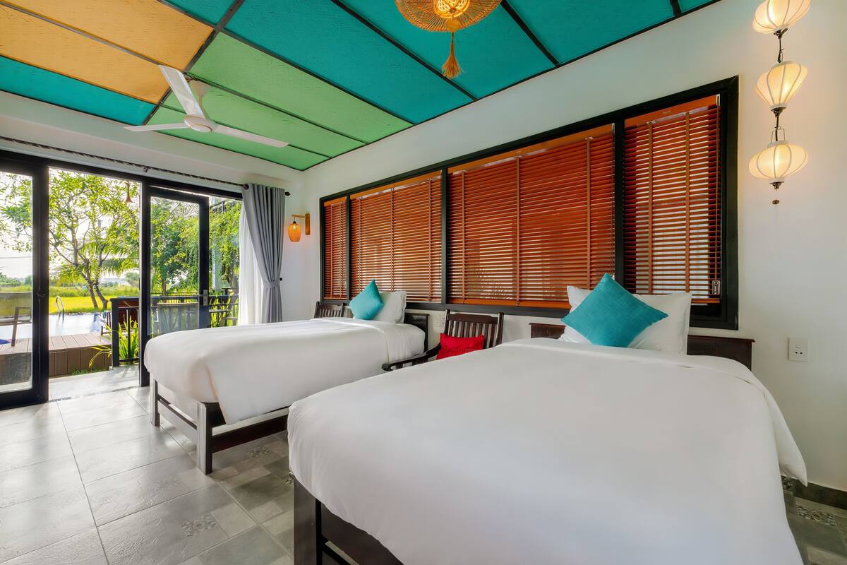 Ein Hotelzimmer mit zwei Betten, einem Deckenventilator, großen Fenstern mit Jalousien und Blick ins Freie.