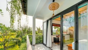 A balcony with a view of a lush green landscape, a sliding glass door leading to a bedroom with a bed and a small table, and a ceiling fan. - Sunkissed Paddy Hoi An Villa (Da Nang)