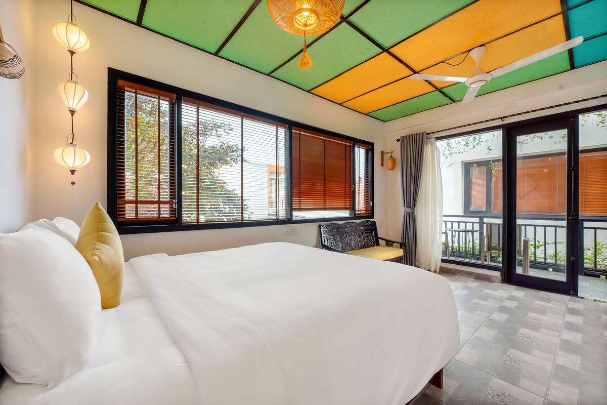 Ein Schlafzimmer mit Bett, Fenster mit Jalousien, Deckenventilator und Blick auf den Balkon.