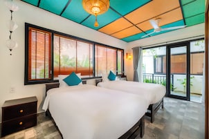 Quarto de hotel com duas camas, ventilador de teto e janelas amplas com persianas.