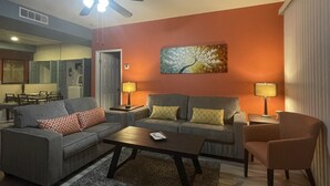 A living room with a grey sofa, a coffee table, and a chair. There is a ceiling fan, a painting on the wall, and a lamp on a side table.