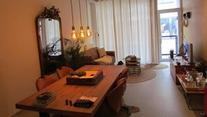 Una sala de estar moderna con una mesa de comedor de madera, un sofá y un espejo grande. Hay luces colgantes y vistas al exterior a través de amplias ventanas.