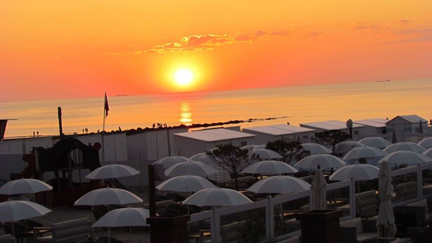 Un hotel frente a la playa con sombrillas y sillas, vista al atardecer y mar en calma.