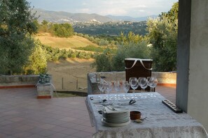 Un espace de restauration en plein air avec une table dressée pour six personnes, offrant une vue panoramique sur un paysage pittoresque composé de collines et d’arbres.