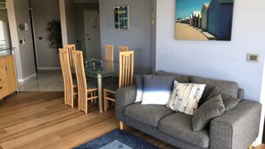 Een woonkamer met een grijze bank, houten stoelen, een glazen eettafel en een blauw vloerkleed.