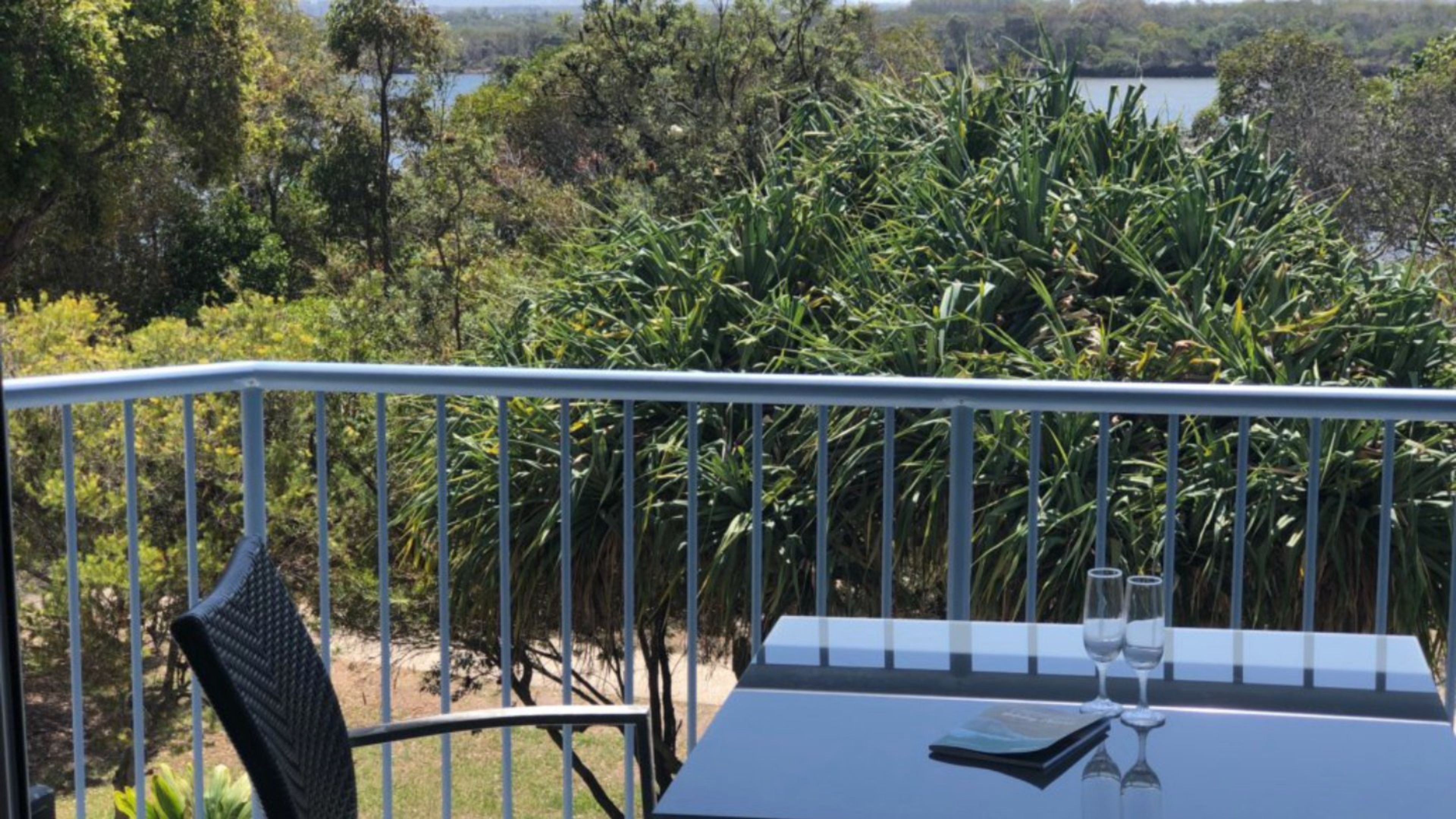 Un balcon avec une table, deux verres et un menu, donnant sur une forêt dense et une étendue d’eau.
