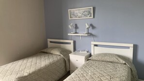Een slaapkamer met twee eenpersoonsbedden, een nachtkastje, een klein ladeblok en een wandplank met twee lampen.