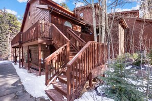 A wooden cabin with a staircase leading to a porch, surrounded by trees and snow.