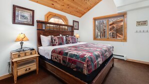 Une chambre avec un plafond en bois, un lit recouvert d’une courtepointe à motifs, une table de chevet, une fenêtre donnant sur un chalet, et un panneau accroché au mur.