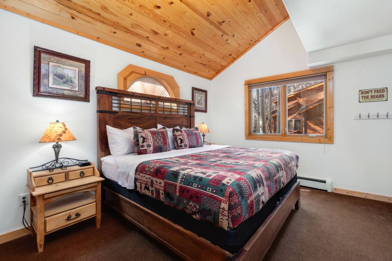 A bedroom with a wooden ceiling, a bed with a patterned quilt, a nightstand with a lamp, and a window with a view of trees.