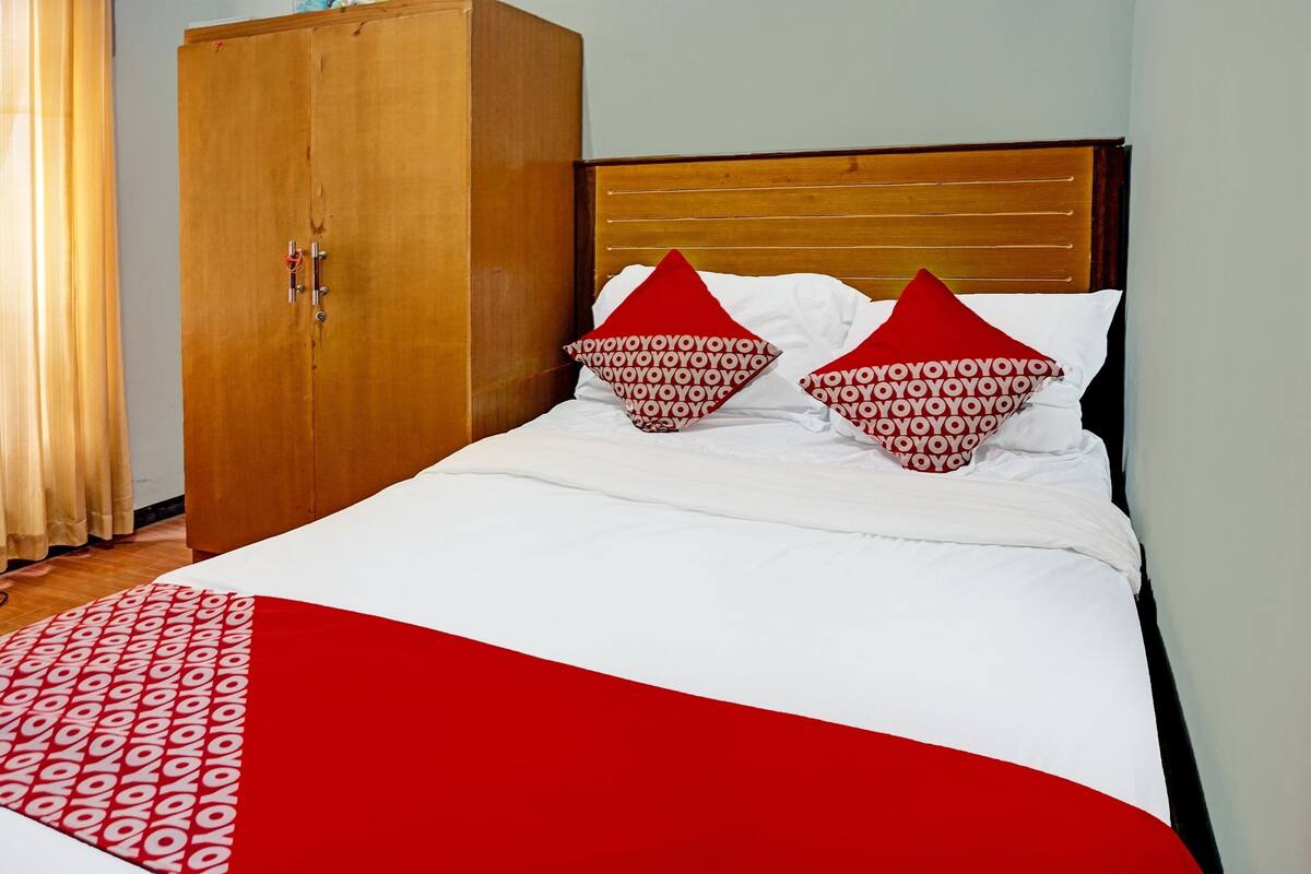 Una cama con ropa de cama blanca, almohadillas rojas, un armario de madera y una colcha estampada en rojo y blanco.