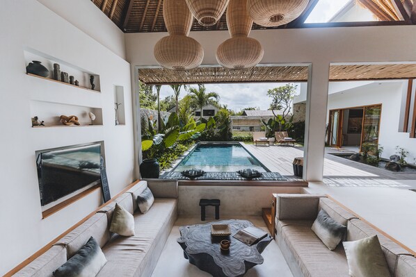 Relax in a spacious living area with a large flat-screen TV, comfortable seating, and a view of the lush garden and pool.