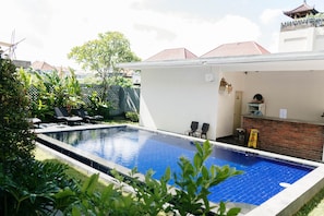 A swimming pool with a tiled deck, surrounded by greenery and a brick wall.