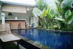 A pool area with a wooden lounge chair, a brick wall, and various tropical plants.