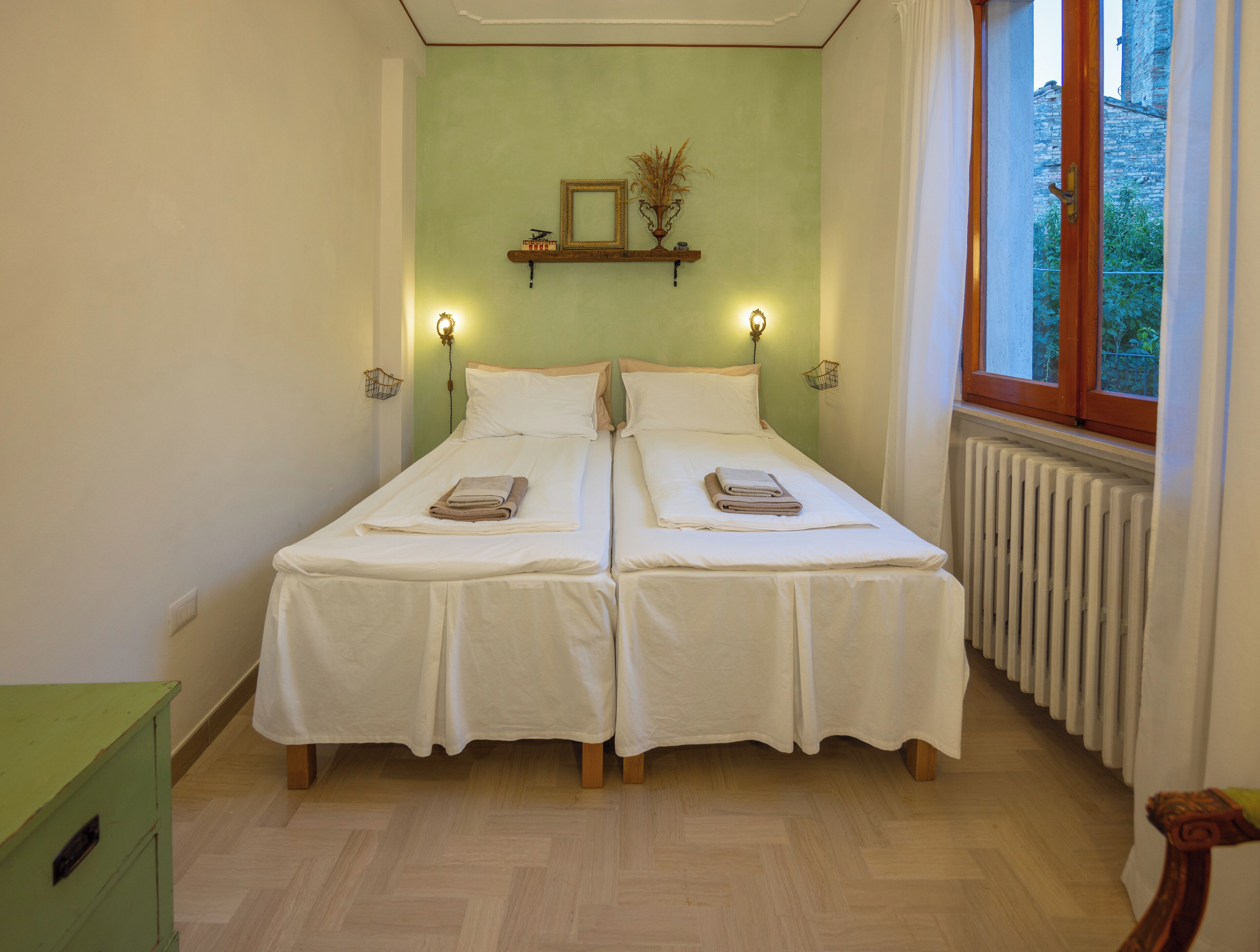 Quarto duplo com duas camas, um radiador, janela com cortinas e prateleira na parede com espelho e objetos decorativos.