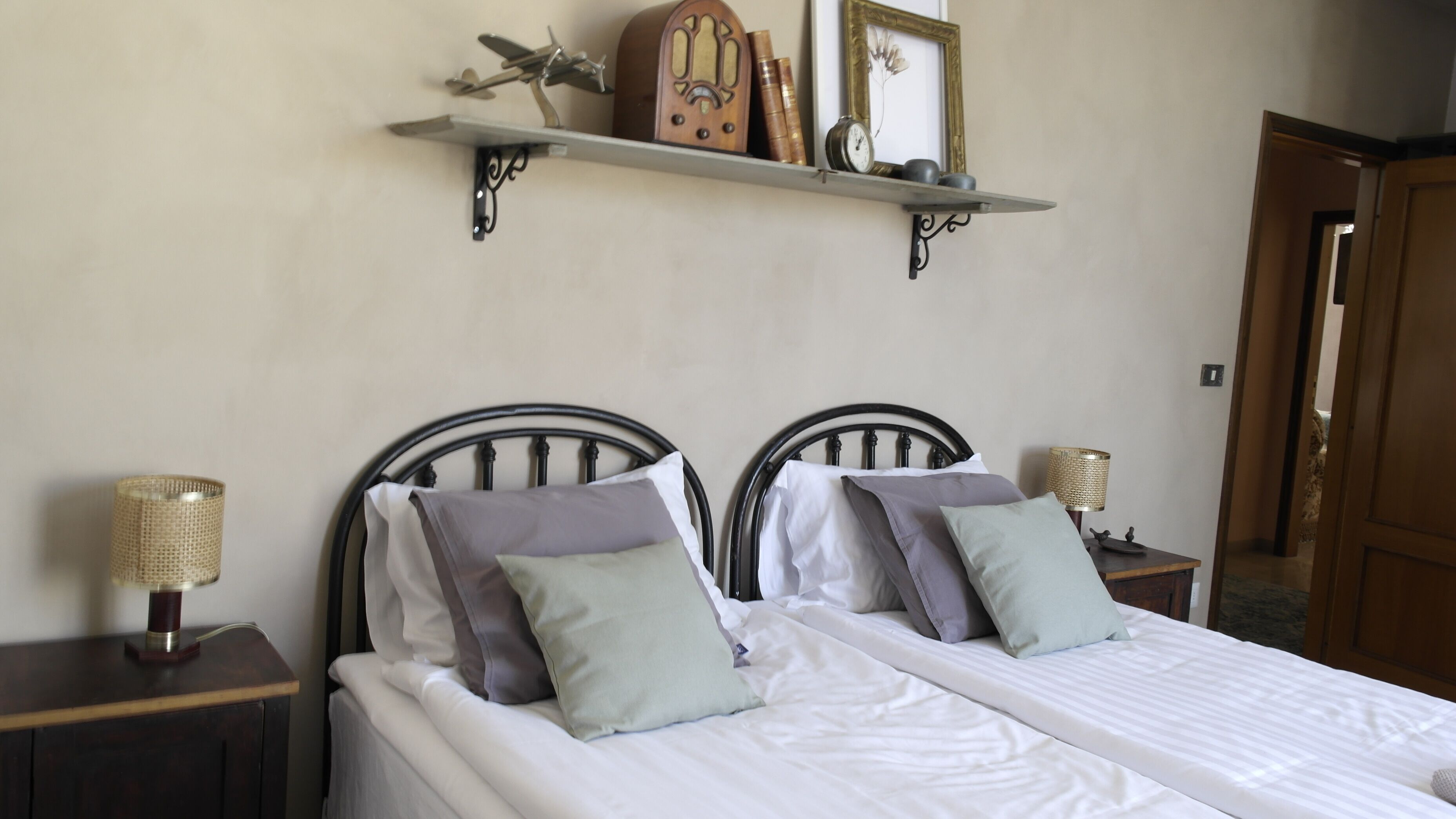 Un lit double avec une literie blanche, deux tables de chevet avec des lampes, une étagère avec des objets décoratifs et une porte en arrière-plan.