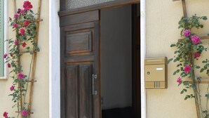 A wooden door with a textured upper section, flanked by potted plants with pink flowers. A mailbox is mounted on the wall to the right. - Casa Agello B&B (Ripatransone)