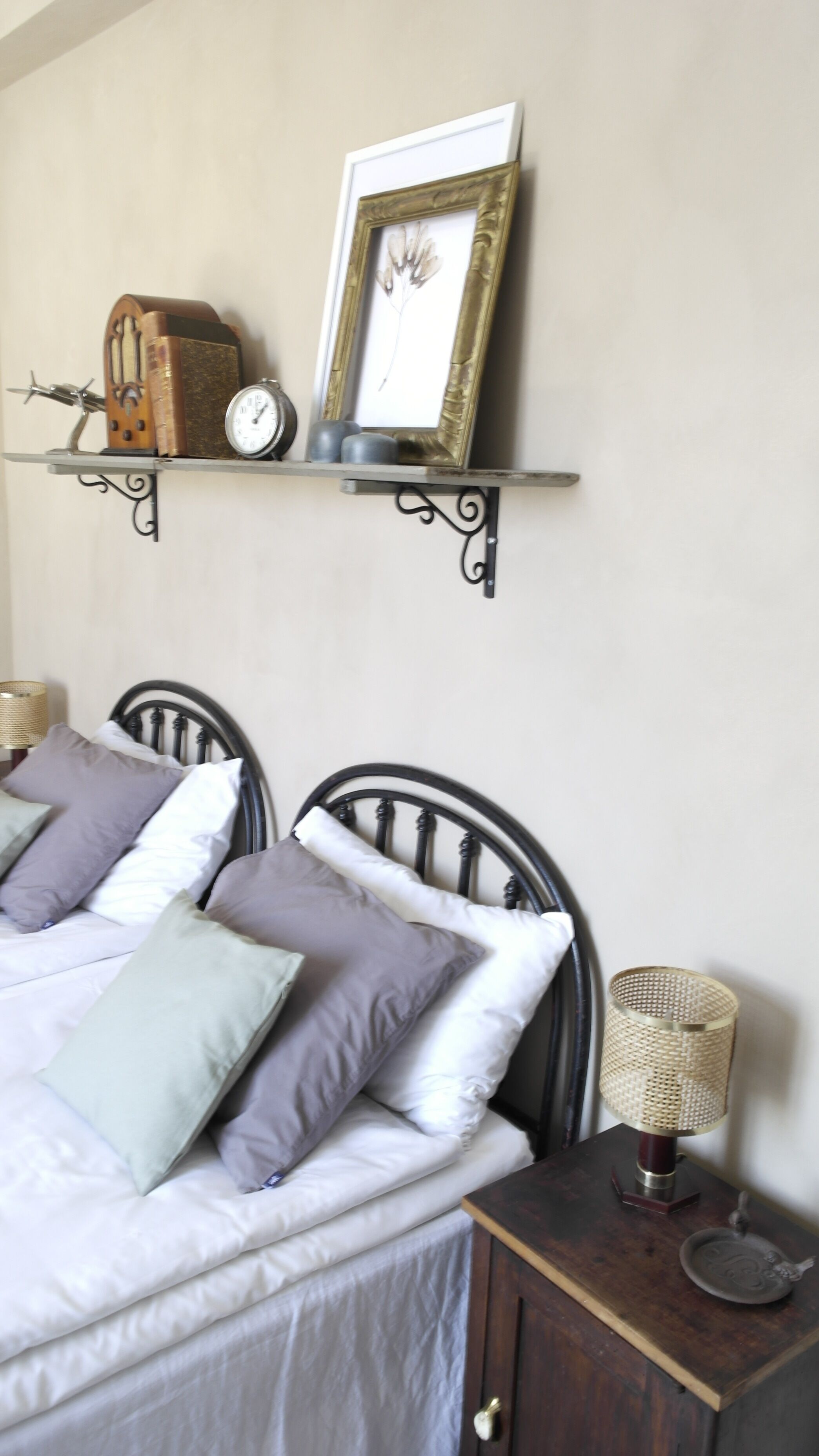 Un letto con testiera, cuscini, un comodino con lampada e una mensola con orologio e oggetti decorativi.