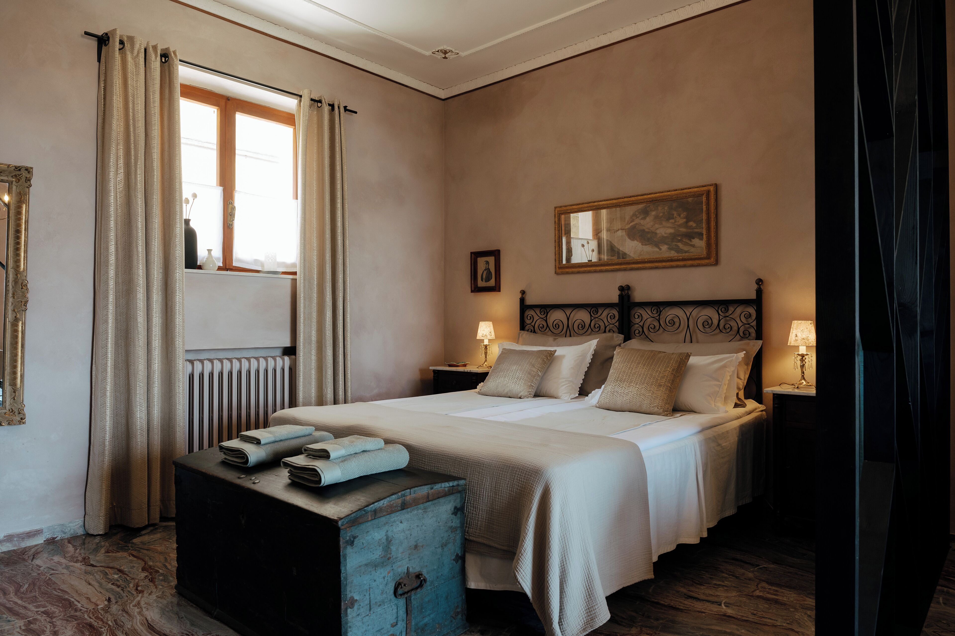 A bedroom with a large bed, two bedside tables with lamps, a dresser, a radiator, and a window with curtains.