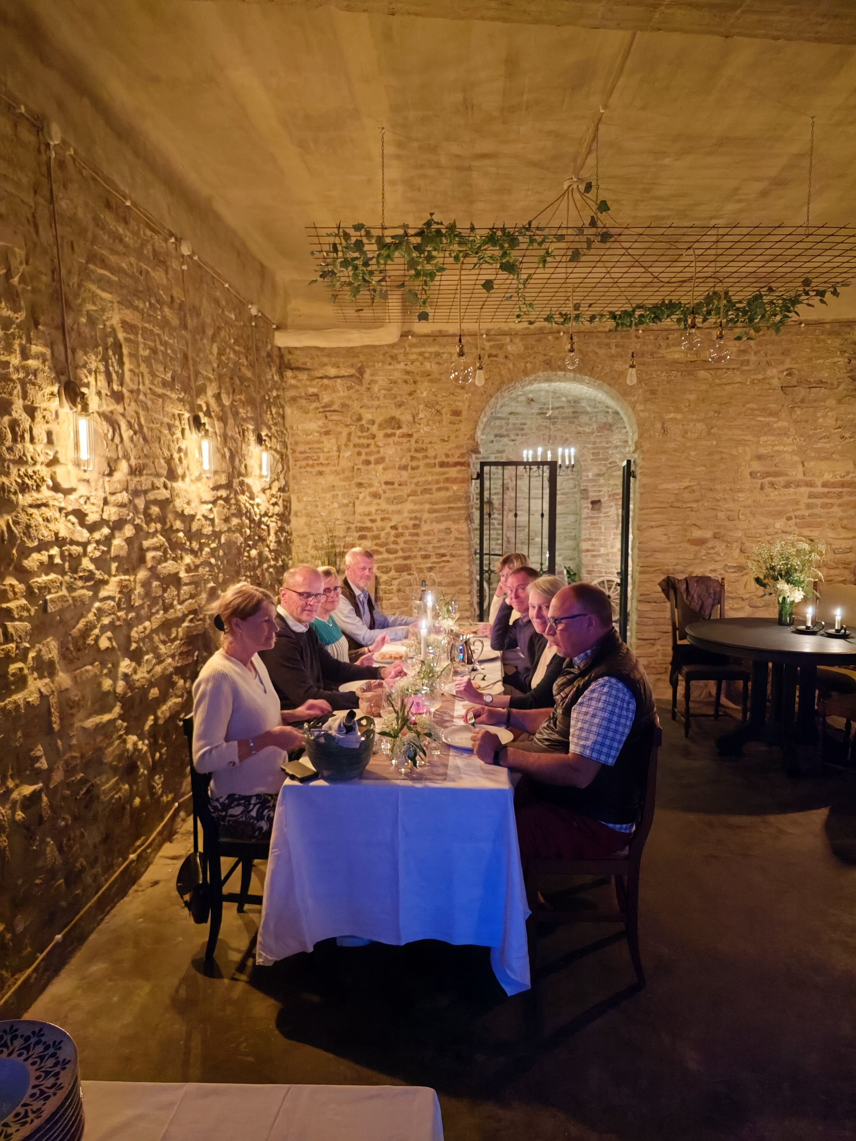 Un groupe de personnes est assis à une table dans une pièce aux murs de pierre, éclairée par des lampes suspendues et un lustre.