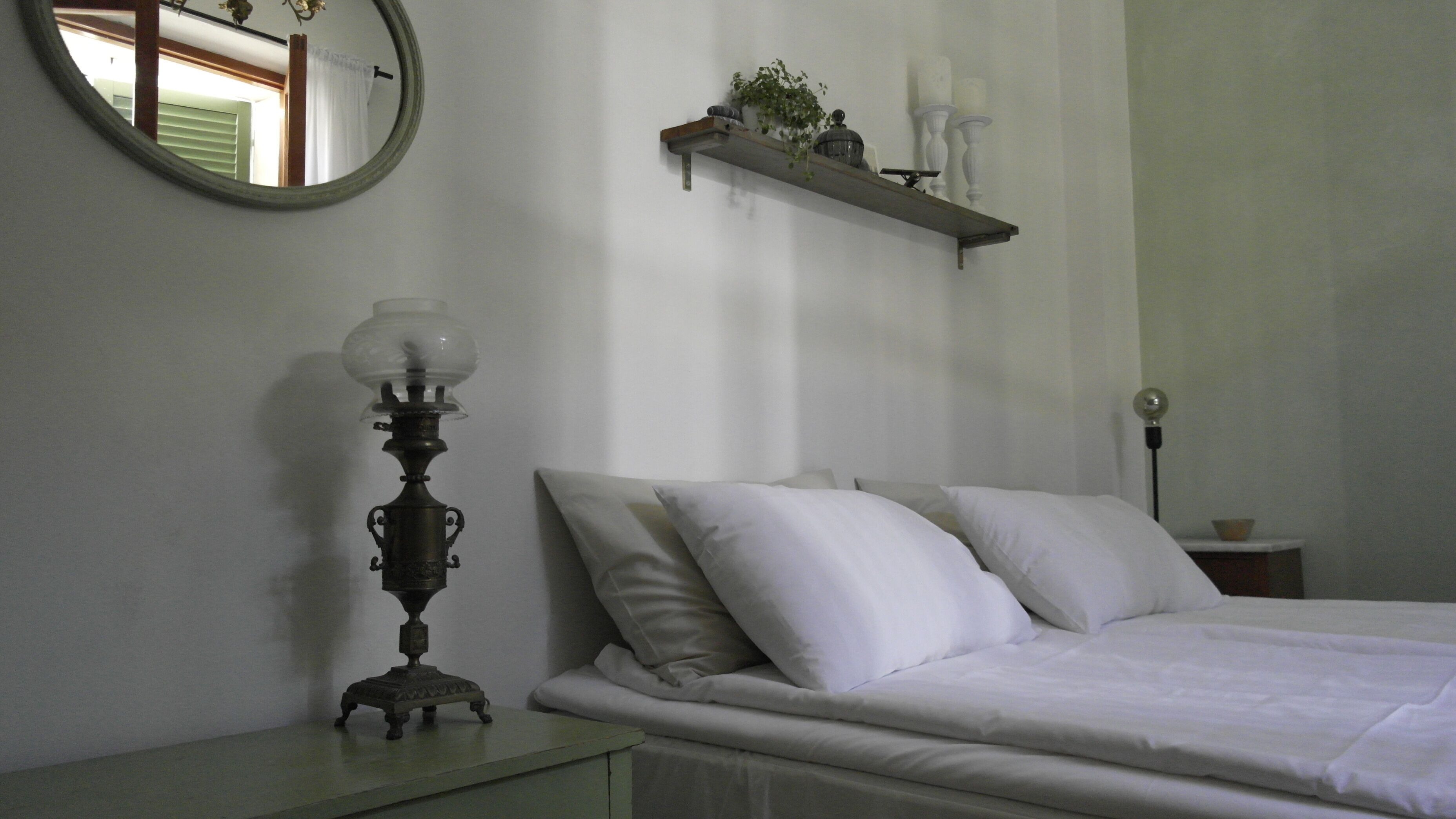 A bedroom with a bed, a nightstand, a mirror, a shelf with candles, and a wall-mounted lamp.