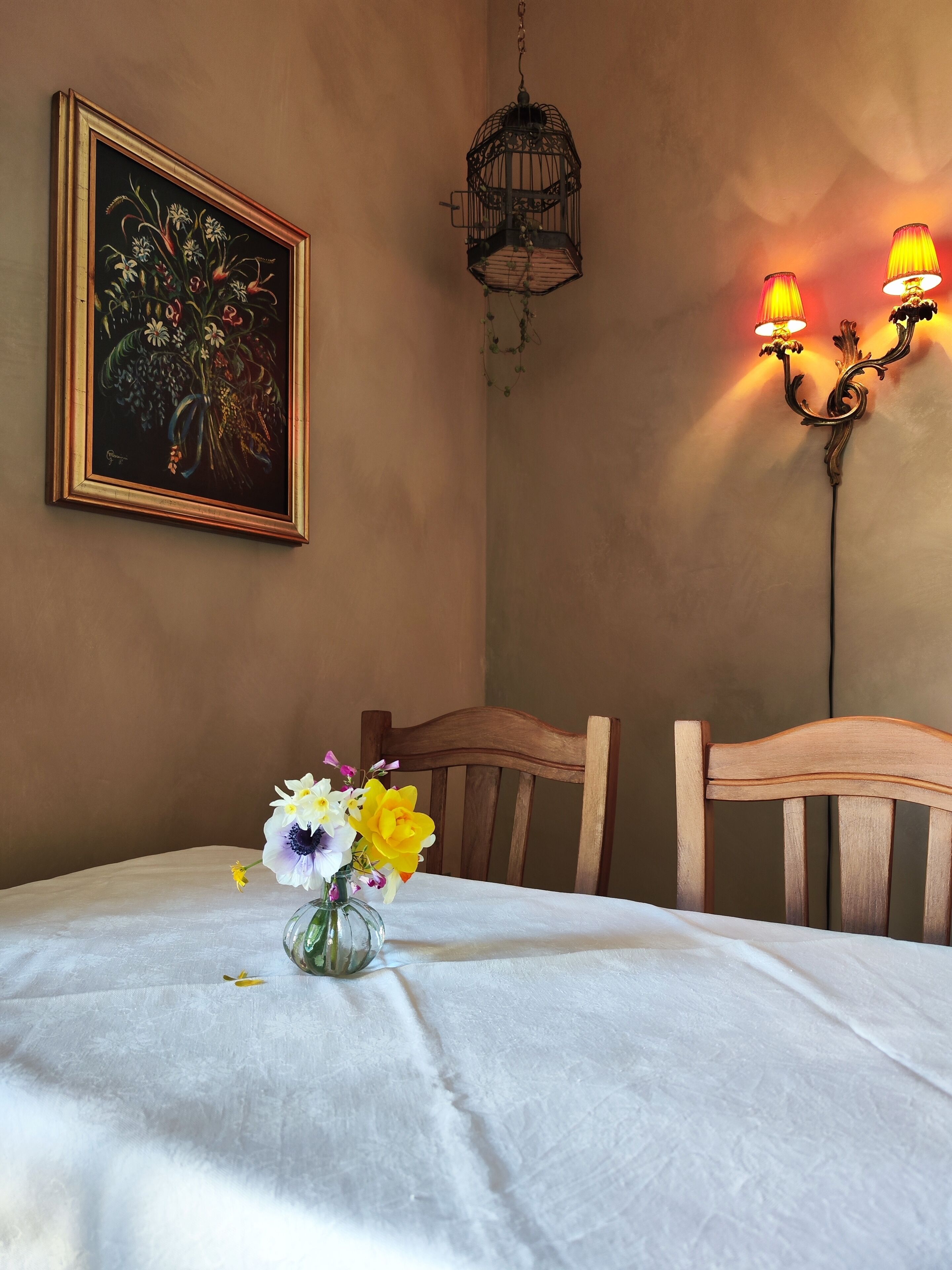 A dining area with a table set for two, a framed painting on the wall, a hanging light fixture, and a wall-mounted lamp.