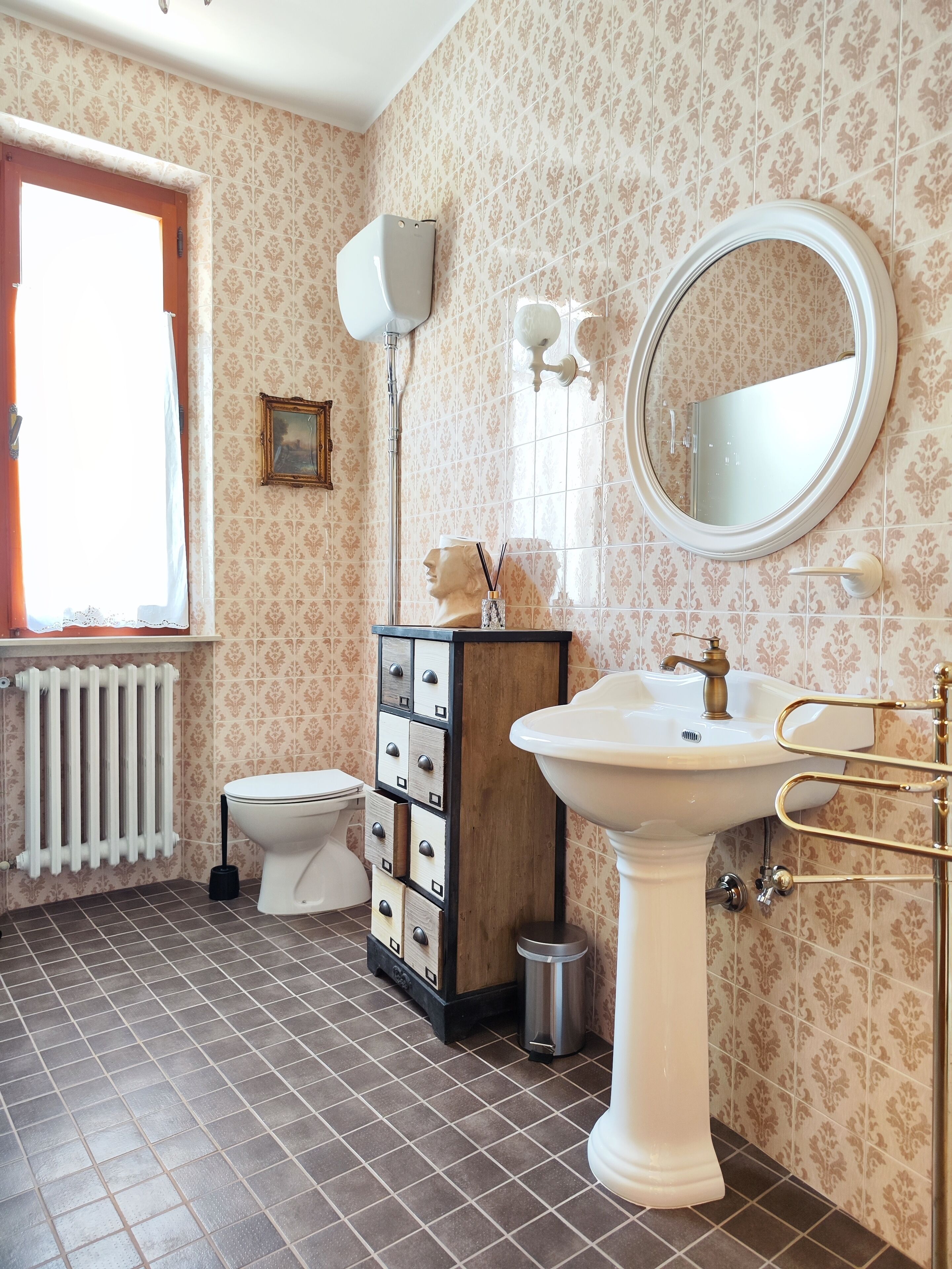 A bathroom with a toilet, sink, and mirror. There is a radiator, a trash bin, and a cabinet with drawers.