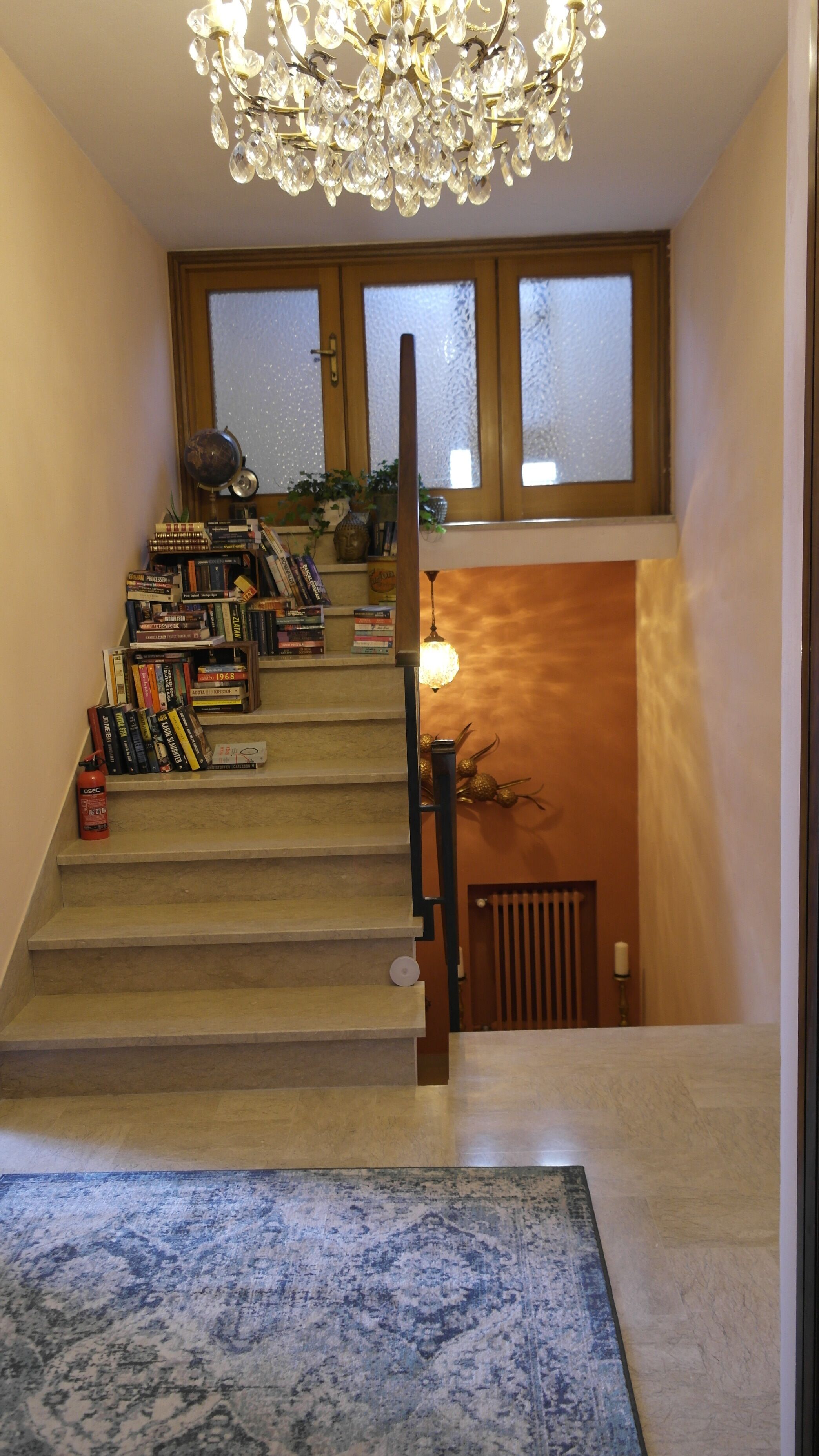 A staircase with a chandelier, bookshelves, and a view of the outside through frosted windows.