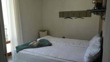 Un letto rifatto con cura, una coperta verde e asciugamani arrotolati, un comodino con una lampada e un attaccapanni a parete con grucce.