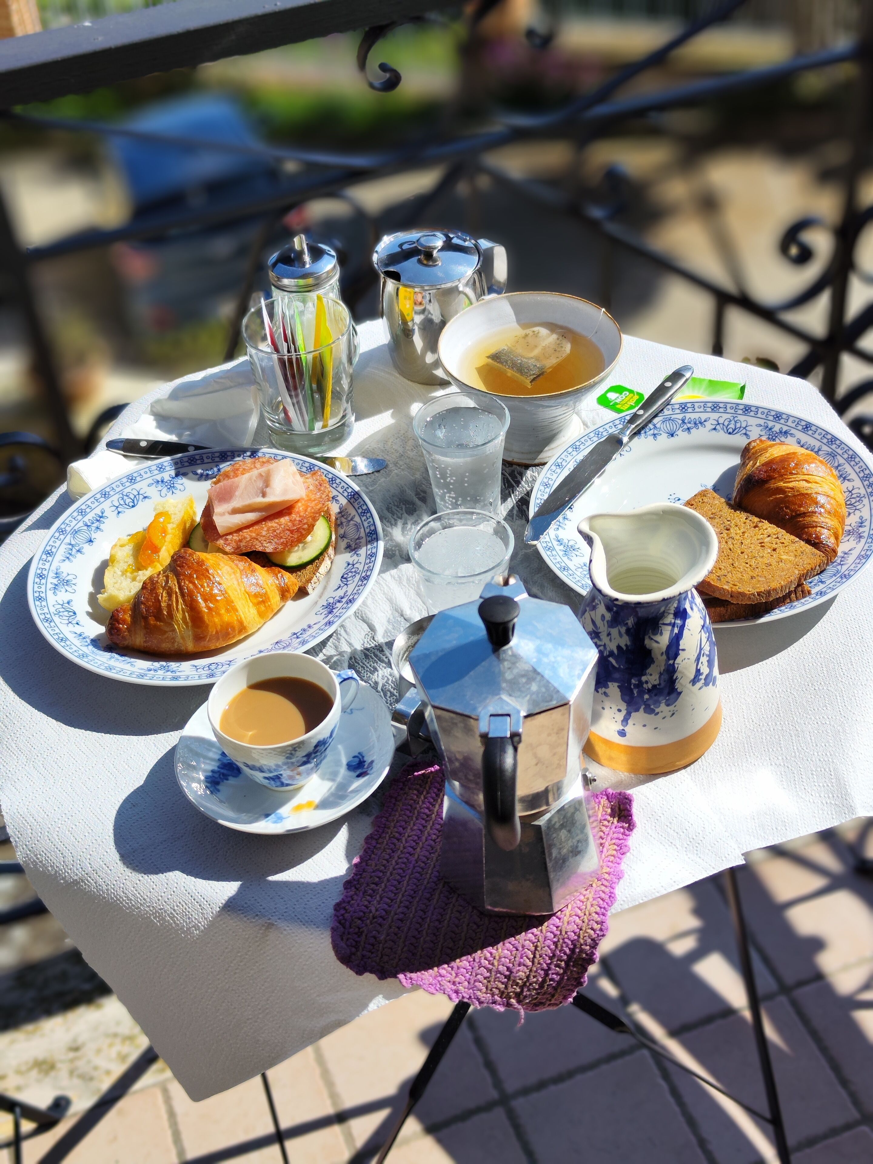 A table set with a croissant, ham, cheese, and bread, along with cups of coffee and tea, on an outdoor table.