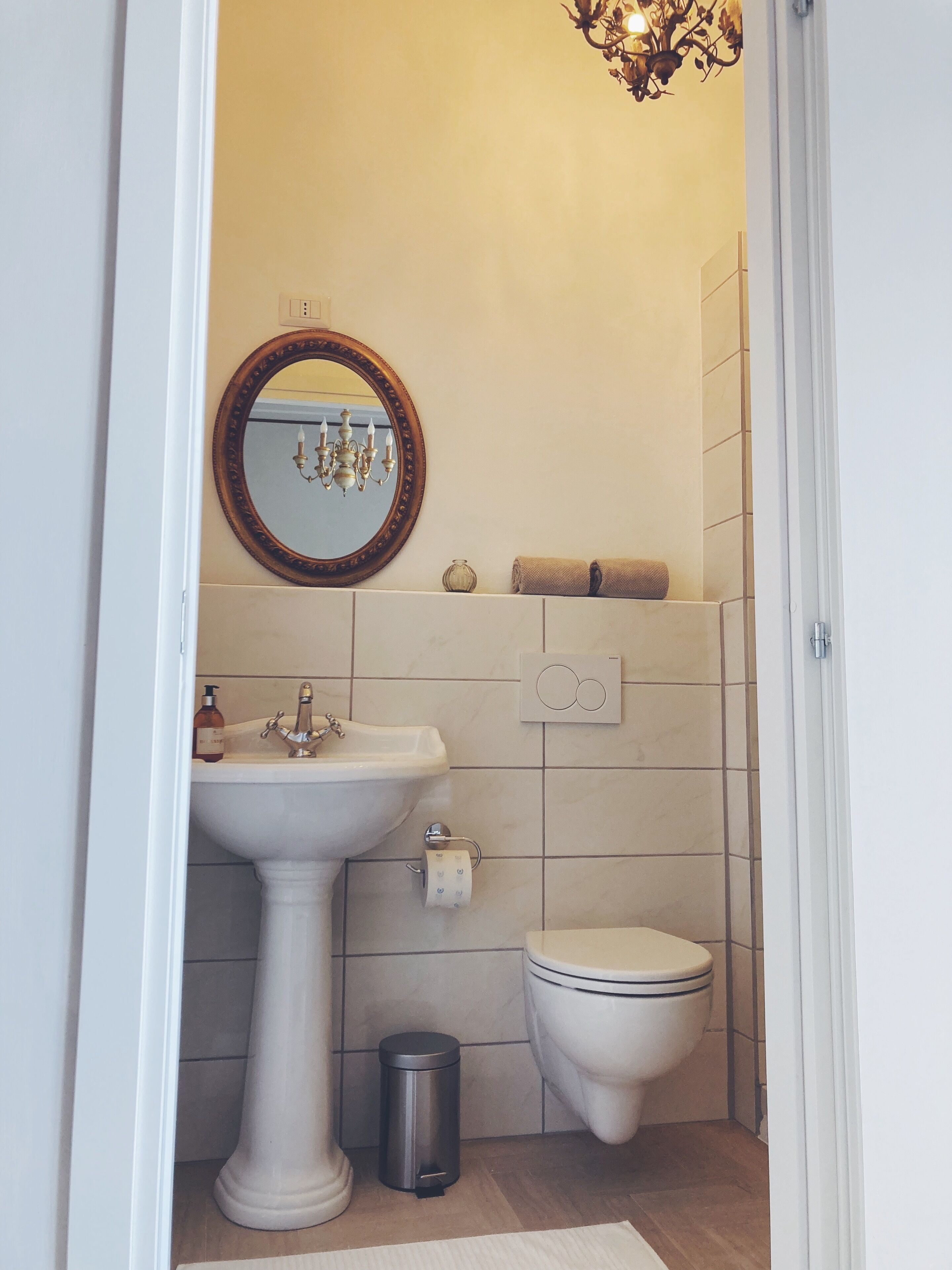 Un baño pequeño con un lavamanos pedestal, un inodoro, un cubo de basura, una toalla y un candelabro.