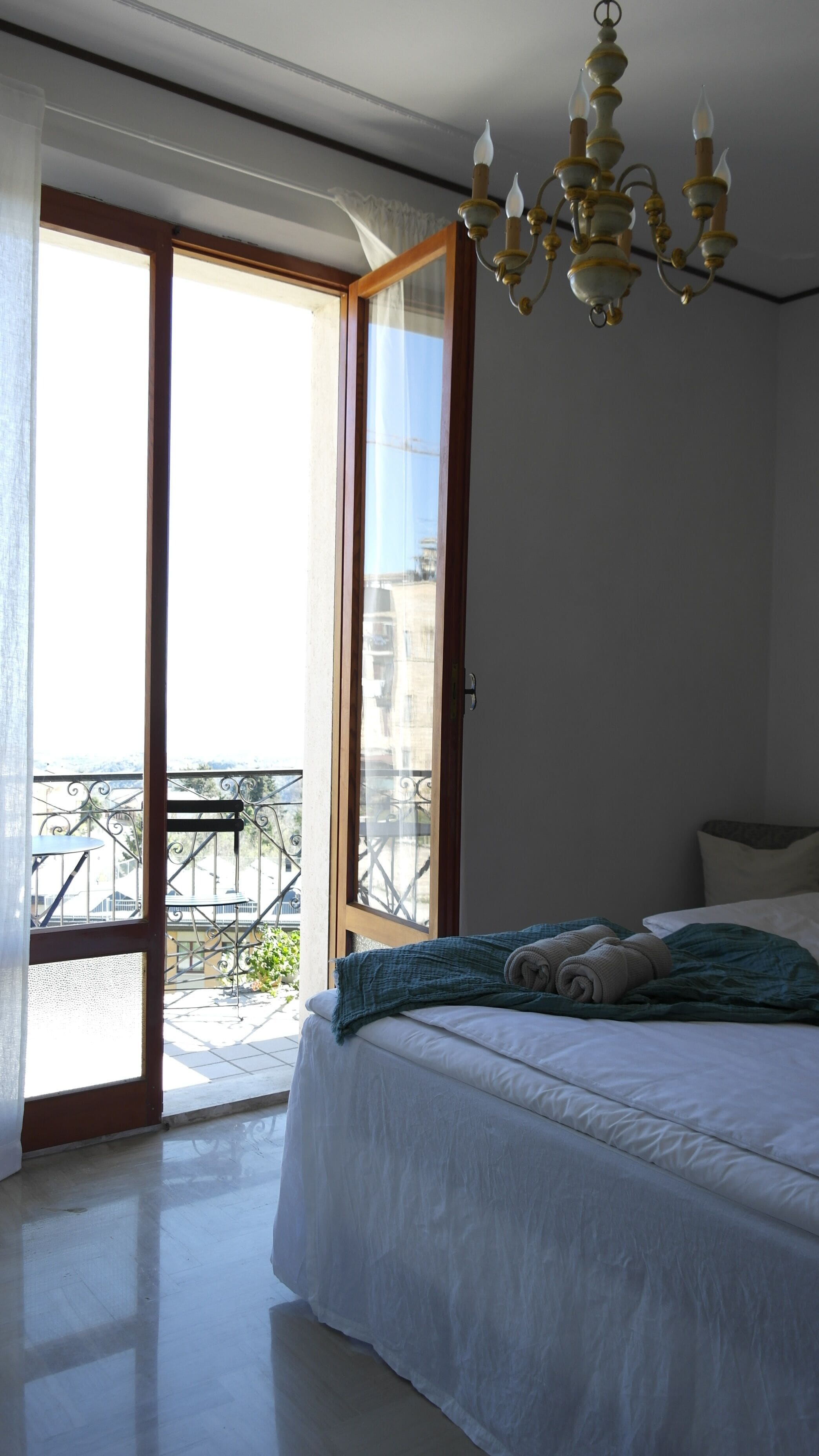 Ein Schlafzimmer mit Bett, einer Chandelier, einem Balkon mit Tisch und Stühlen und Blick auf Gebäude.