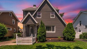 Une maison à deux étages avec un porche à l’avant, une clôture en piquets et deux arbres à feuilles persistantes.