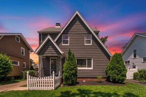 A two-story house with a front porch, a picket fence, and two evergreen trees.