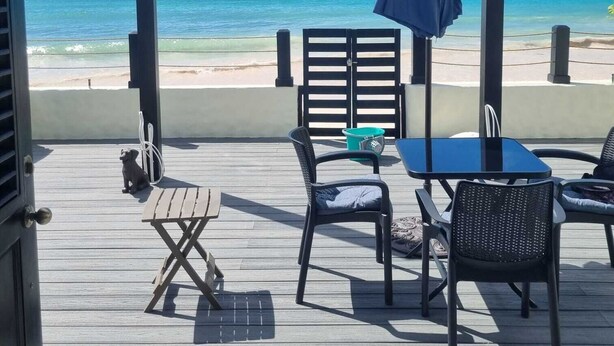 Een terras buiten met een tafel en stoelen, met uitzicht op een strand met helderblauw water en een zandstrand.