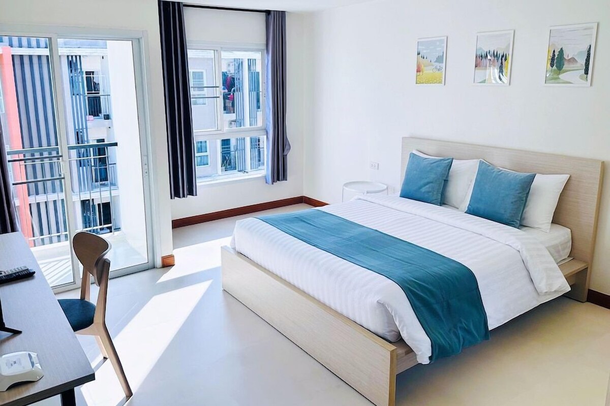Une chambre d’hôtel avec un lit, un bureau et une chaise. Il y a une fenêtre avec des rideaux, et des œuvres d’art sont accrochées au mur.