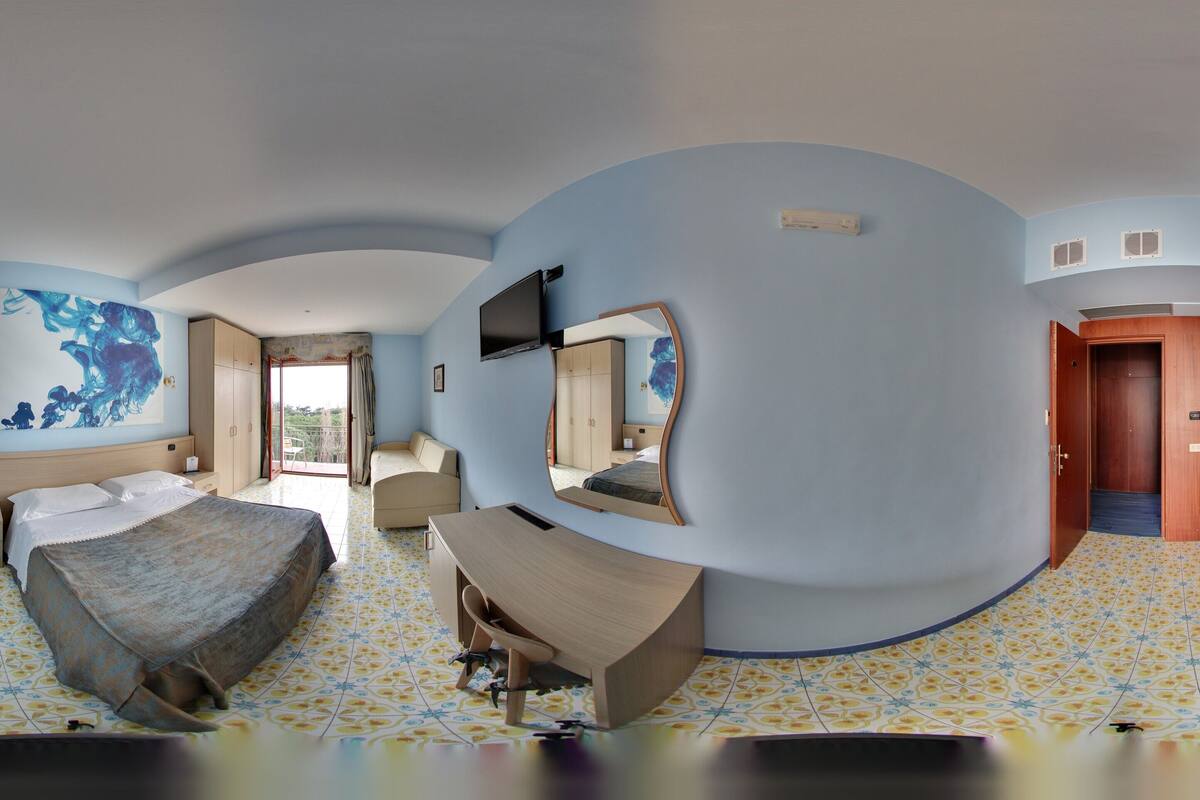 A bedroom with a bed, a desk, a chair, a large mirror, and a painting on the wall.