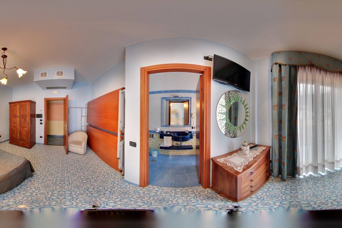 A bedroom with a bed, a wooden wardrobe, a small table, a mirror, and a bathroom visible through an open door.
