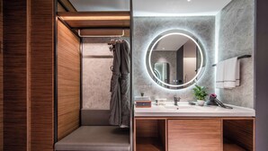 A modern bathroom with a wooden vanity, a round mirror with underglow lighting, a towel rack, and a built-in shoe storage area.