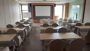 A classroom with rows of desks and chairs, a projector screen, and a large window.