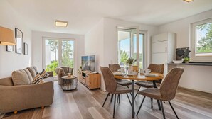 A modern living room with a sofa, a coffee table, and a dining area with chairs and a table. There is a TV on a stand and a window with a view of greenery. - Hochwertiges Ferienappartement für bis zu 4 Personen in Ruhiger Wohngegend an de (Zirchow)