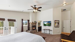 A bedroom with a large bed, a fireplace, a ceiling fan, a TV, and a door leading to a balcony.