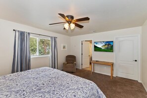 Un dormitorio con una cama, un ventilador de techo, una silla, un televisor y una puerta que da a otra habitación.