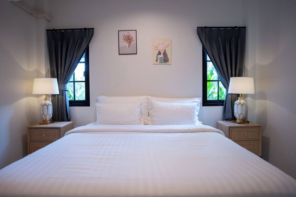 A neatly made bed with white linens, two bedside tables with lamps, and two windows with curtains.