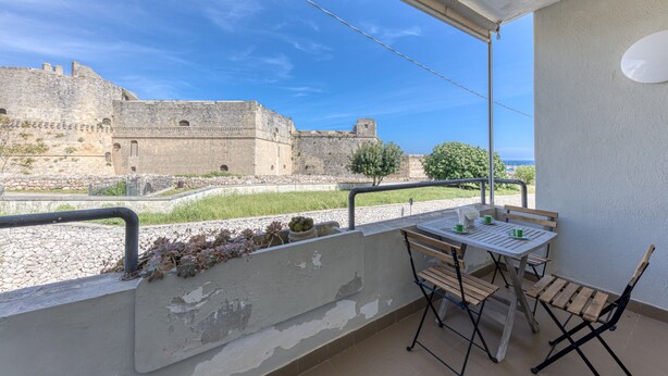 En balkon med et bord og stole, udsigt over en historisk bygning i sten og et græsareal.