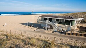 Um edifício à beira-mar com uma escadaria que leva a um deck, uma praia de areia e o oceano ao fundo.