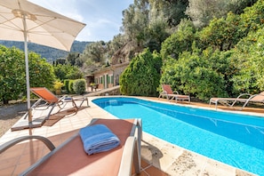 A swimming pool with lounge chairs and an umbrella, surrounded by greenery and a building.