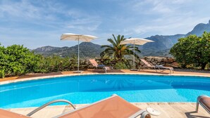 Una piscina con sillas de descanso y sombrillas, rodeada de vegetación y montañas al fondo.