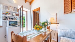 Un comedor con una mesa de madera, sillas, un cuenco de fruta, un jarrón con flores, una lámpara y un refrigerador con utensilios de cocina.